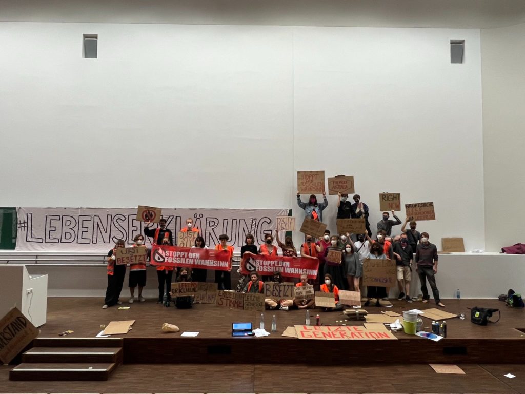 Gruppenfoto auf der Bühne eines Universitäts-Hörsaals. Die Tafel wurde mit einem Banner mit der Aufschrift "Lebenserklärung" überhängt. Die Personen halten Plakate hoch.