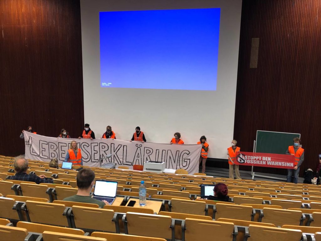 A lecture hall. People wearing high-visibility vests stand on the stage and hold up a banner with the inscription "Declaration of Life".