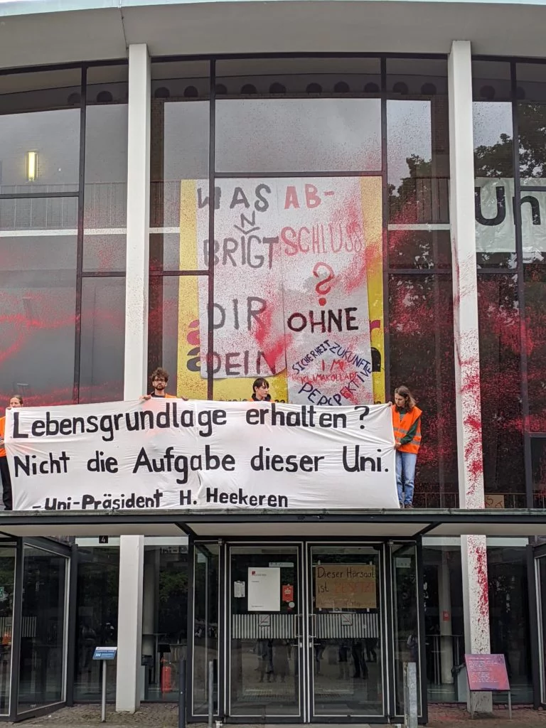 People hold a banner with the inscription "Preserve livelihoods? Not the task of this university. - University President H. Heekeren". The building behind them is colored with red-orange paint.