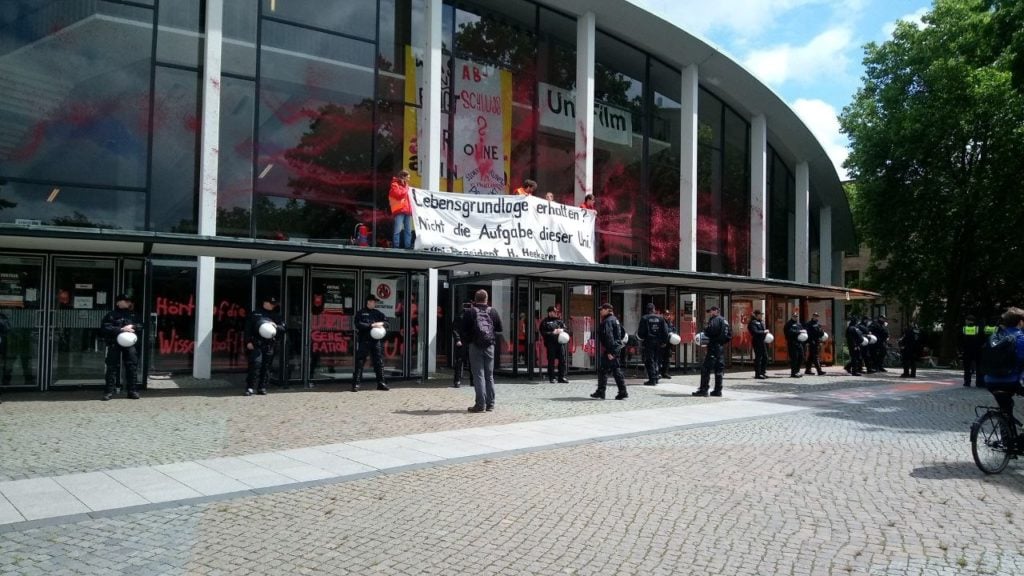 The orange-red colored auditorium is surrounded by police. People hold up a banner on the entrance canopy with the inscription "Preserve livelihoods? Not the task of this university. - University President H. Heekeren".