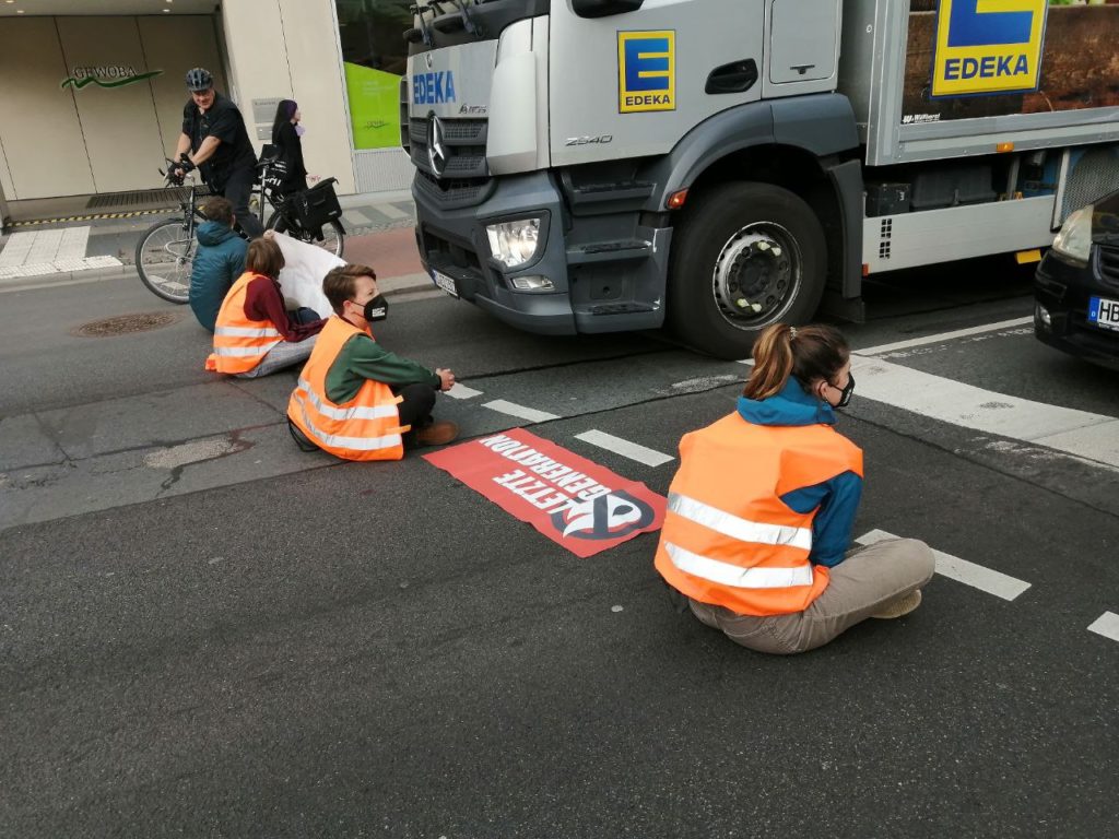 Several people are sitting on a street in high-visibility vests with their backs to the viewer. Between them lies a banner with the inscription "Last Generation". There is a truck and a car in front of them.