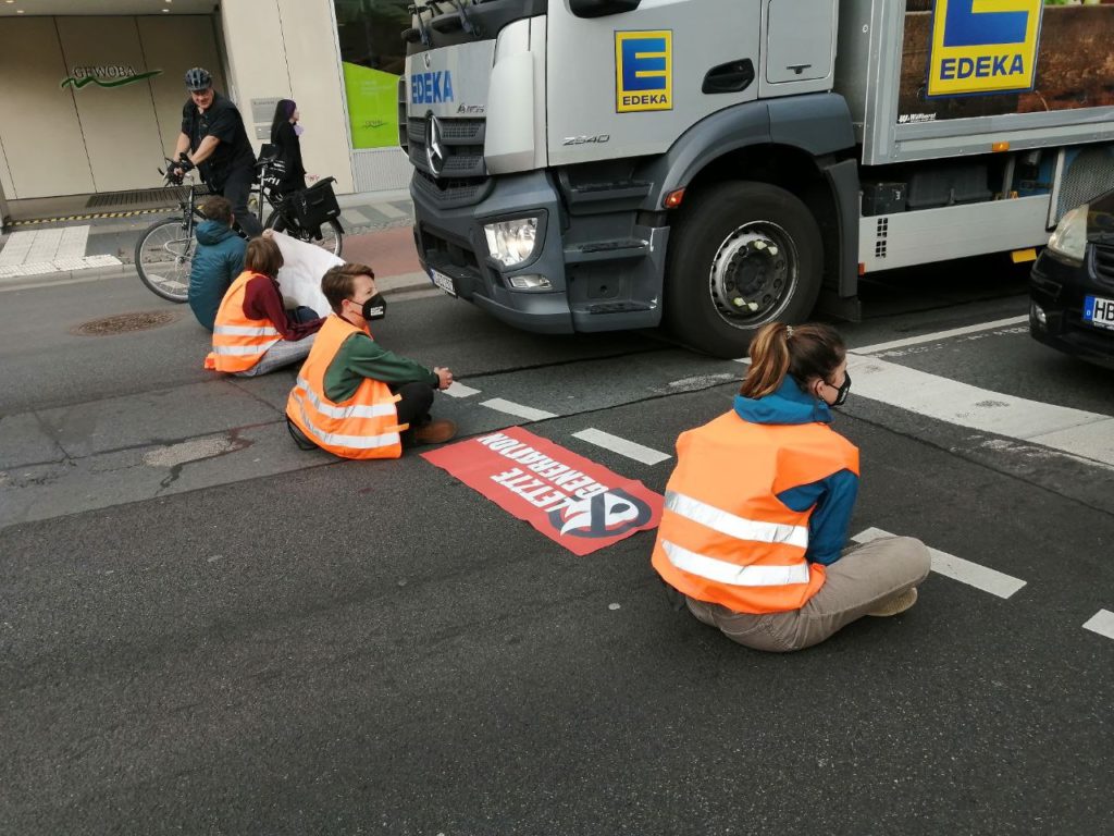 Mehrere Menschen sitzen mit dem Rücken zum Betrachte in Warnwesten auf einer Straße. Zwischen ihnen liegt ein Banner mit der Aufschrift "Letzte Generation". Vor ihnen steht ein LKW und ein Auto.