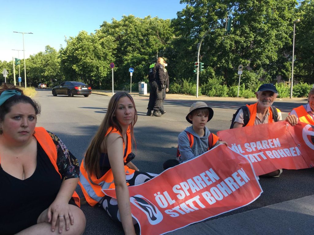 Four people block the street sitting next to each other with banners and high-visibility vests. The middle two are children.