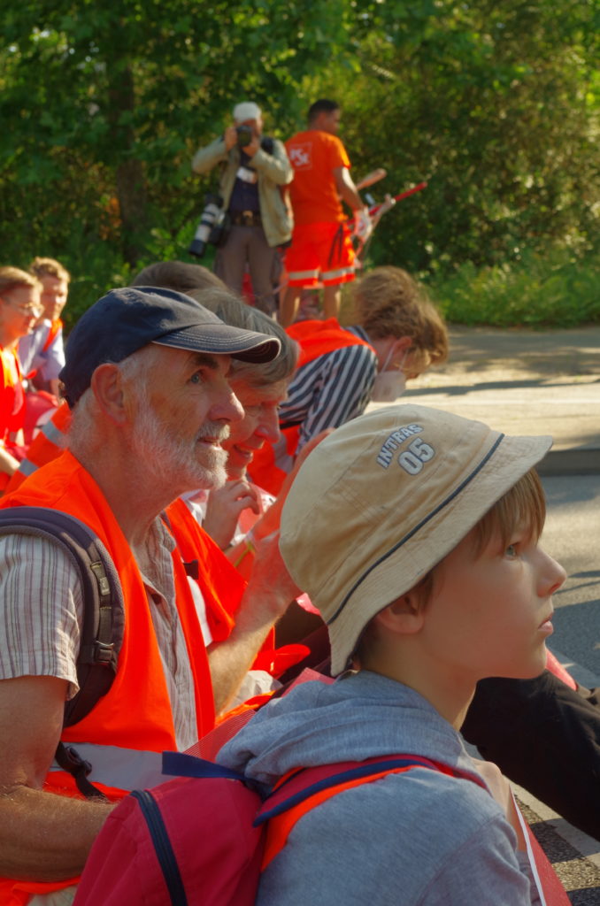 Several people wearing high-visibility vests sit in a row next to each other on the street. They look to the right. At the front is a child, also wearing a safety vest.