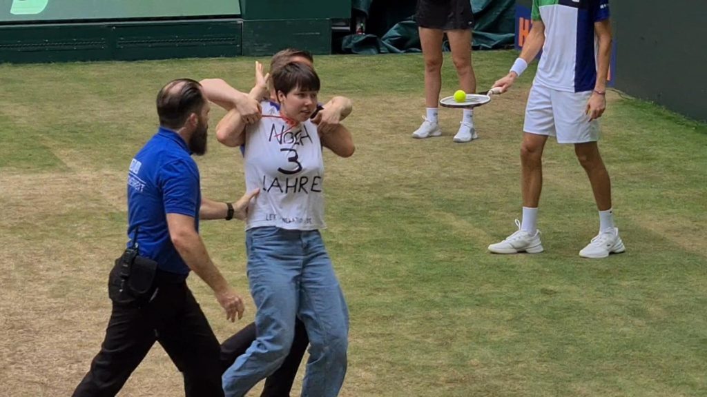 A person is taken away from a tennis court by security. She wears a shirt that says “3 more years.”