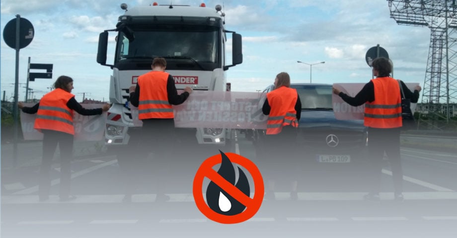 4 Menschen stehen mit Bannern auf der StraĂe. Sie stehen in einer Linie mit dem RĂŒcken zum Betrachter. Sie tragen schwarze Klamotten unter ihren Warnwesten. Ihnen gegenĂŒber stehen Autos.