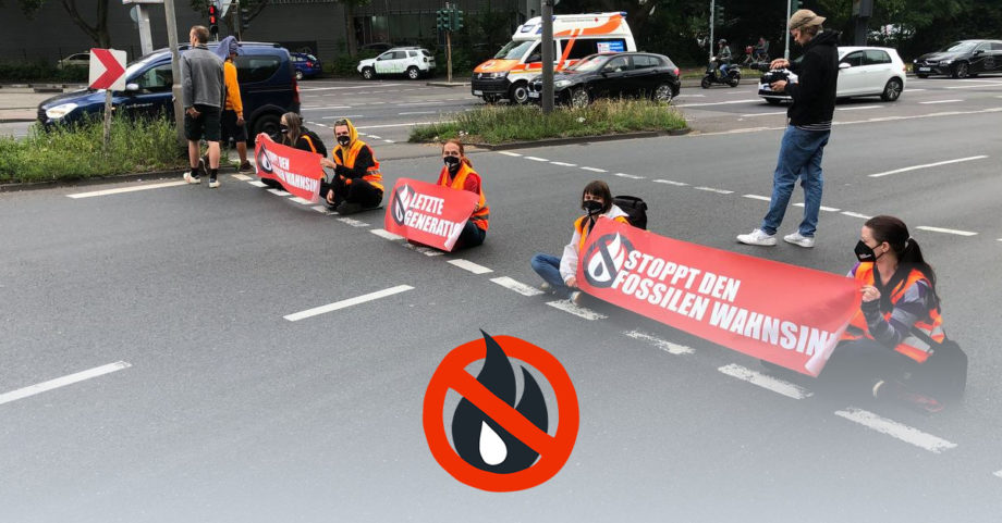 Mehrere Menschen in Warnwesten sitzen an einer roten FuĂgĂ€ngerampel mit Bannern auf der StraĂe. Auf den Bannern steht "Stoppt den fossilen Wahnsinn" oder "Letzte Generation".