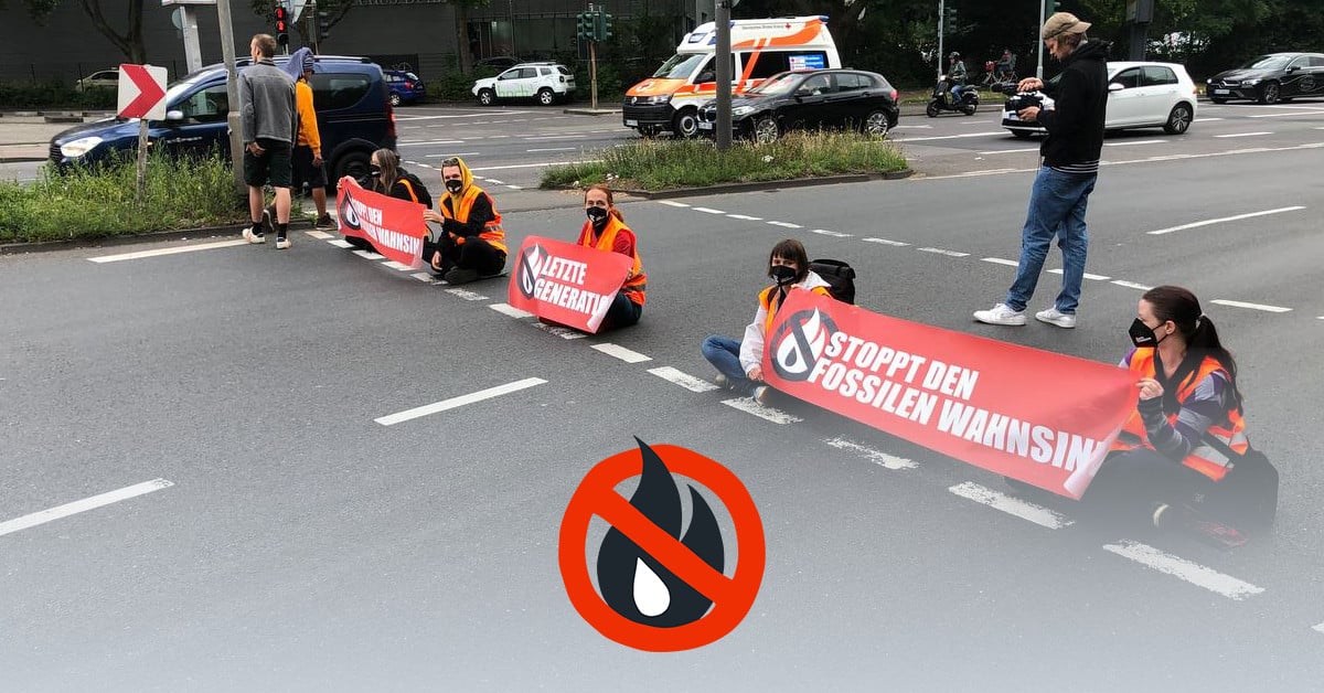 Several people in high-visibility vests sit on the street at a red pedestrian light with banners. The banners say “Stop fossil madness” or “Last Generation”.