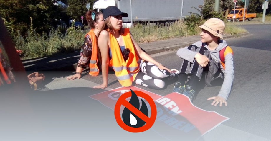 Zwei Kinder sitzen in Warnwesten auf der StraĂe. Vor ihnen liegt ein Banner mit der Aufschrift "Ăl sparen statt bohren". Jeweils eine ihrer HĂ€nde ist auf die StraĂe geklebt..