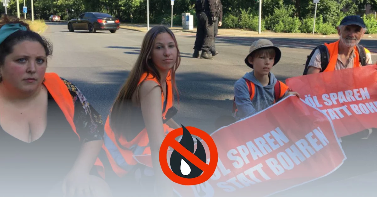 Four people block the street sitting next to each other with banners and high-visibility vests. The middle two are children.