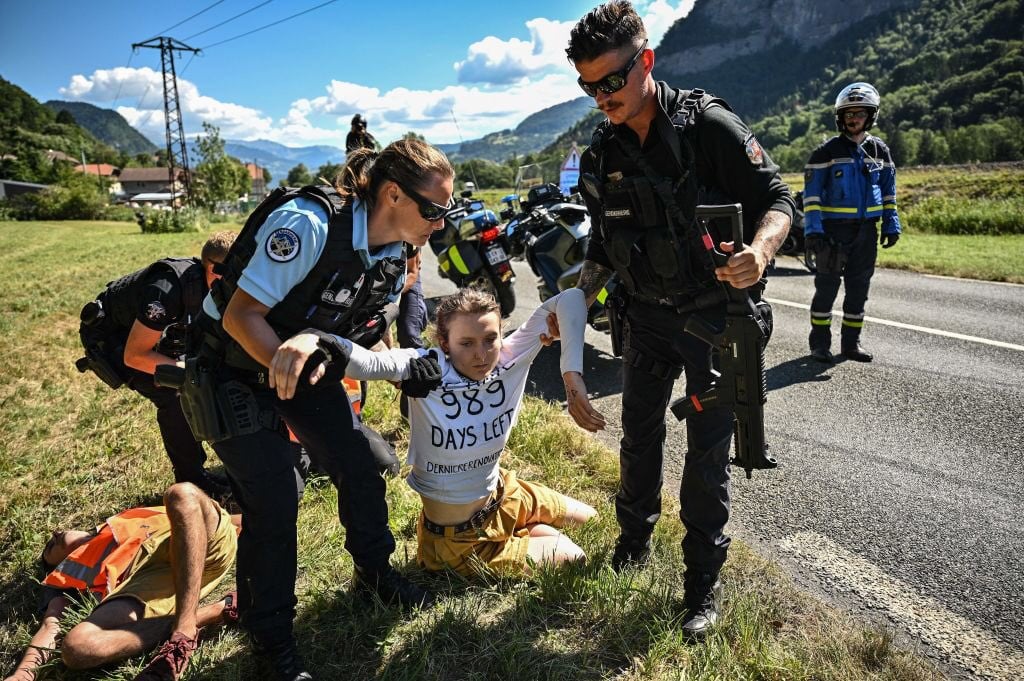 A person gets pulled away from a street by the police. She wears a shirt on which is written "We have 989 days left" and "dernierrenovation.fr".