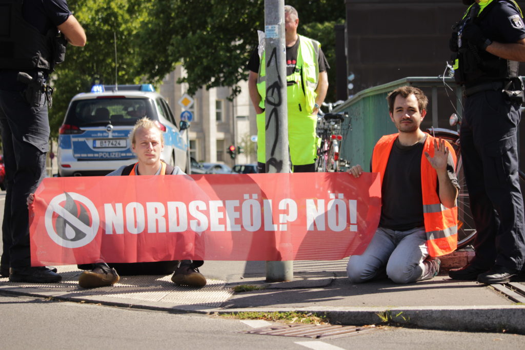 Two people in high-visibility vests block the street. They hold a banner that says "North Sea oil? Nope!" in the hands.