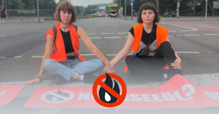 Zwei Bürgerinnen sitzen nebeneinander auf der Straße und halten sich an den Händen. Sie tragen Warnwesten und vor ihnen liegt ein Banner mit der Aufschrift "Nordseeöl? Nö!".