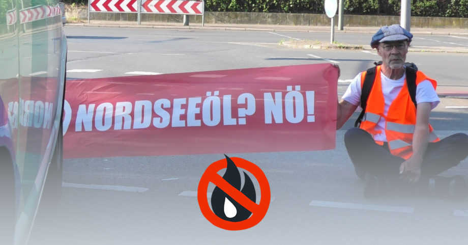 Ein älterer Mann sitzt vor einem Auto auf der Straße. Er sitzt frontal zur Kamera gerichtet. Er trägt eine orangene Warnweste und hält ein Banner mit der Aufschrift "Nordseeöl? Nö!". Foto: Mark Dominik Bürkle