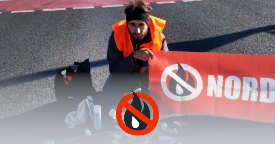 Ein Mensch sitzt auf der Straße. Er hat eine Warnweste an und hält ein Banner in der Hand. Seine andere Hand klebt an der Straße. Vor sich liegt Kleber und ein Schild mit der Aufschrift "Achtung angeklebt".