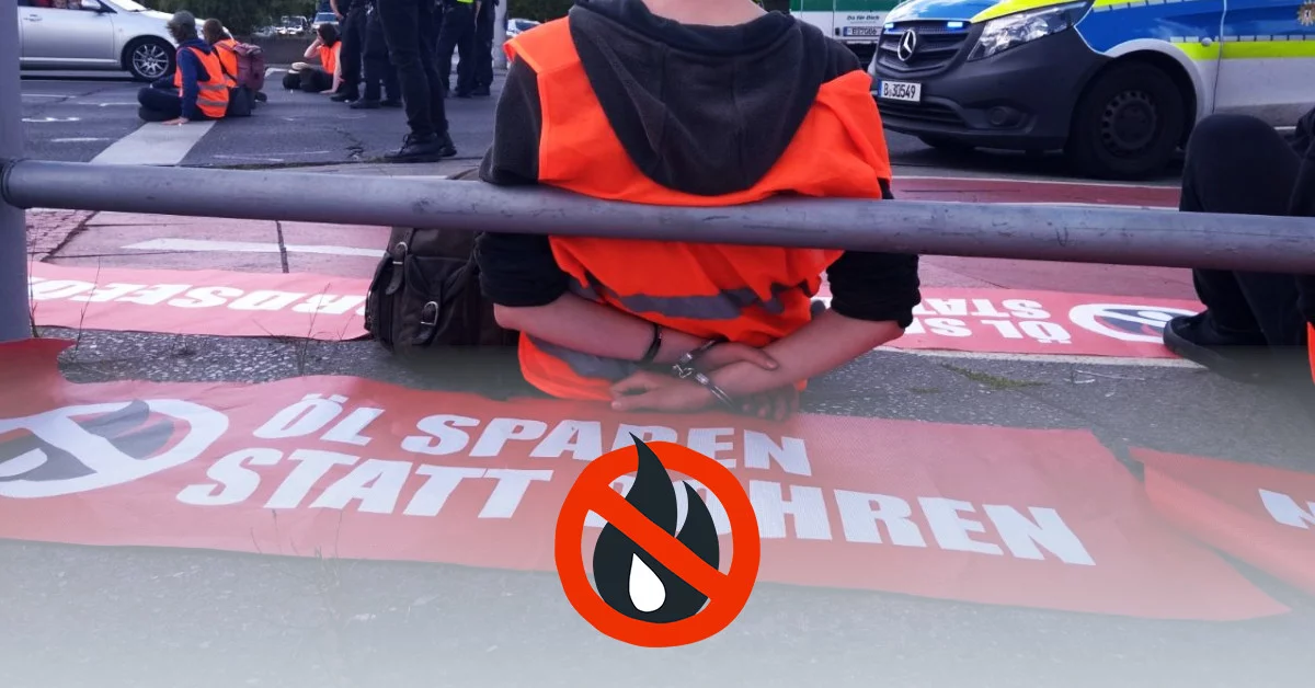 A person sits in handcuffs on the side of the road. The person is wearing a high-visibility vest and around him are banners from the last generation with the inscription “Save oil instead of drilling”. In the background, other people in high-visibility vests are sitting on the street.