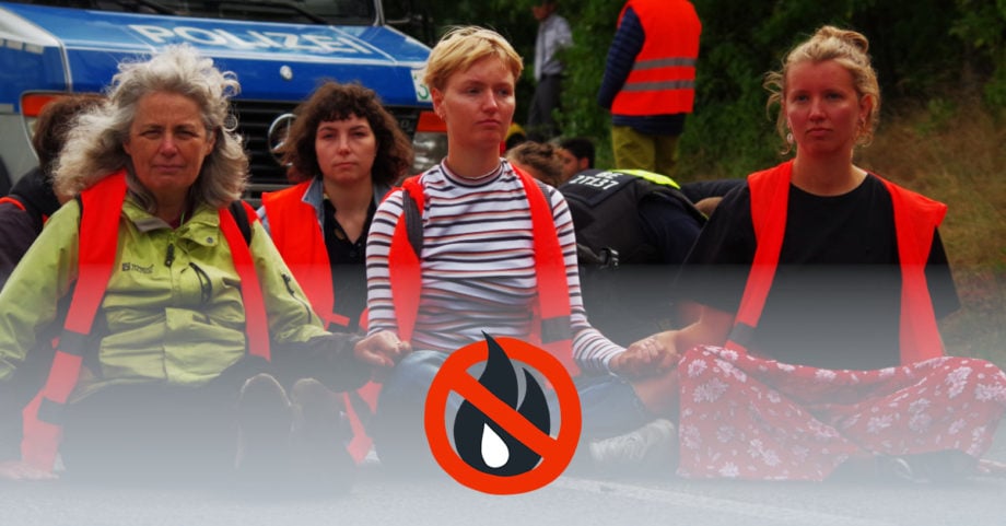 Straßenblockade der Letzten Generation. Die 3 Blockierenden im Vordergrund halten sich friedlich und entschlossen an den Händen während sie in Warnwesten auf der Straße sitzen. Im Hintergrund ist ein Polizeiauto zu erkennen.
