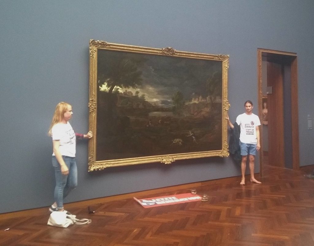 Zwei Menschen kleben mit ihren Händen an dem Gemälde "Gewitterlandschaft mit Pyramus und Thisbe". Sie tragen T-Shirts mit der Aufschrift "Letzte Generation - Stoppt den fossilen Wahnsinn!". Vor ihnen liegt ein Transparent der Letzten Generation.