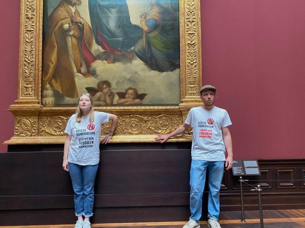 Zwei Menschen kleben mit ihren Händen an dem Gemälde "Sixtinische Madonna". Sie tragen T-Shirts mit der Aufschrift "Letzte Generation - Stoppt den fossilen Wahnsinn!".
