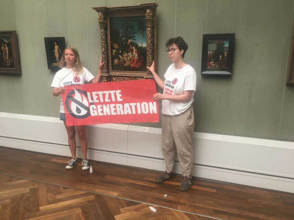 Lina Eichler and Maja Winkelmann each stick their hands on one side of the painting “Rest on the Flight to Egypt” by Lucas Cranach. Between them they hold a banner with the inscription “Last Generation”. Their T-shirts say "Last Generation - Stop the Fossil Madness!".