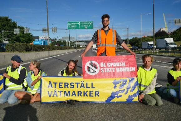 Kevin Hecht steht mit einem Banner mit der Aufschrift "Öl sparen statt bohren" auf der Straße. Um ihn herum sitzen Menschen der “Återställ Våtmarker” ebenfalls mit Warnwesten und Bannern mit dem Organisationsnamen.