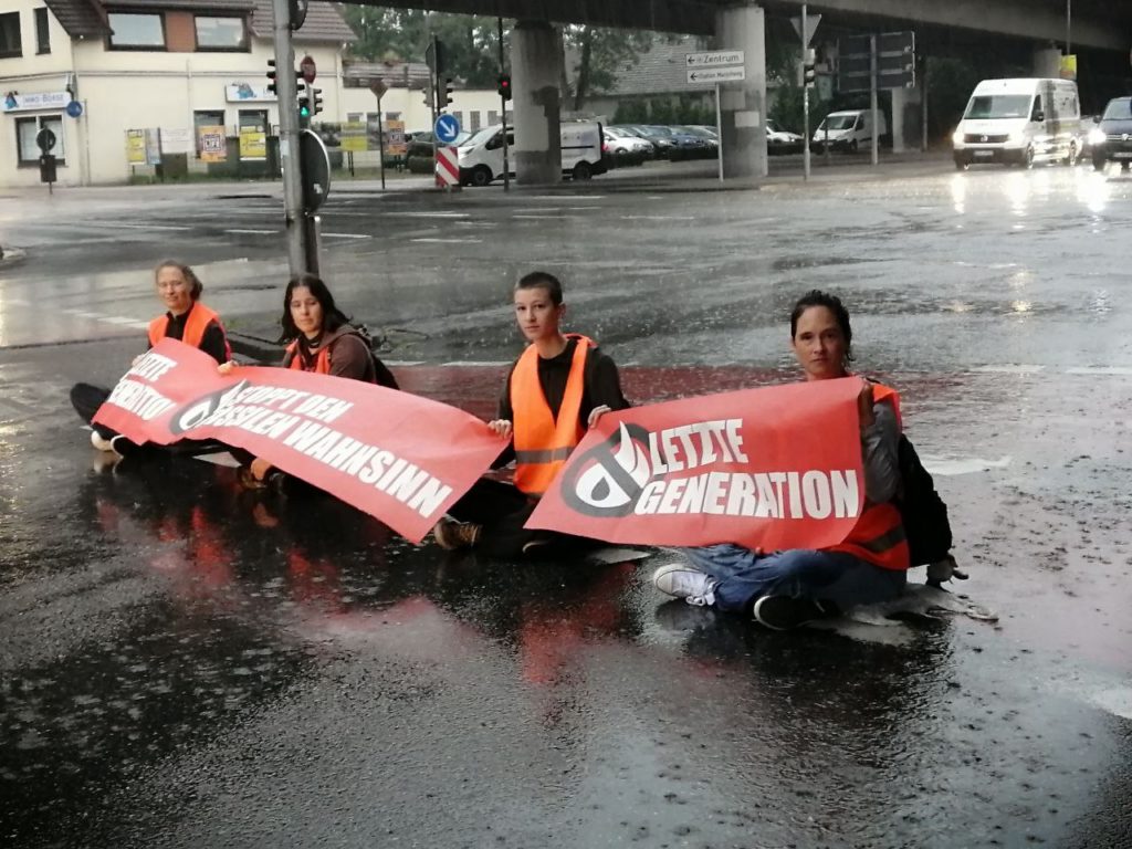 Four people are sitting on the street in the rain wearing high-visibility vests. They hold banners with the inscription “Stop fossil madness” or “Last Generation” in their hands.