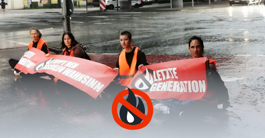 Vier Menschen sitzen im Regen in Warnwesten auf der Straße. Sie halten Banner mit der Aufschrift "Stoppt den fossilen Wahnsinn" oder "Letzte Generation" in den Händen.